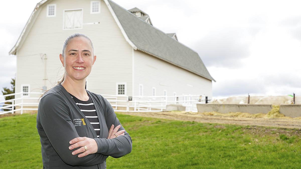 A woman standing in grass field with a tan barn and fencing in the background.