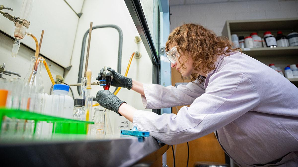 Woman in chemistry lab wearing safety goggles and lab coat examines a beaker.